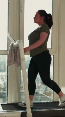 Genç tombul kadın evde koşu bandında yürüyor, kardiyo antrenmanı yapıyor. Evde fitness antrenmanı. Spor ve eğlence konsepti. Dikey video