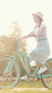 Romantik, hayalperest ve ilham verici mavi elbiseli kadın güneş ışığında klasik geçmişe dönük bisiklete biniyor. Şapkalı ve elbiseli güzel bir bayan güneş ışığında parkta eski bir bisiklete biniyor. Dikey video