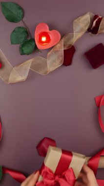 Kadın eli, Sevgililer Günü için paketlenmiş hediye kutusuna kırmızı kurdele bağlıyor. Kadın 14 Şubat için hediye hazırlıyor. Güllü masa, yanan mumlar ve hediyeler. Dikey video