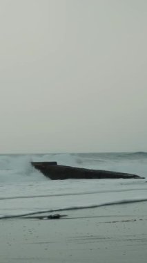 Fırtınalı denizde boş bir iskele. Güçlü okyanus dalgaları kumlu sahile çarpıyor, su sıçrıyor ve beyaz köpük oluşturuyor. Rüzgarlı bir günde büyük dalgalarla dramatik bir deniz. Dikey video