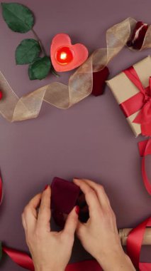 Çiçekler, mumlar, hediyeler ve şömineler eşliğinde Aziz Sevgililer Günü arkaplanındaki nişan yüzüğünün olduğu küçük kırmızı kutuyu eller açıyor. Dikey video