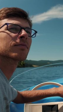 Portrait of young male in glasses sailing on electric boat along mountain lake. Cheerful man floating on yacht at sunny day during summer vacation. Vertical video