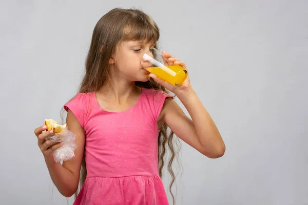 Uma menina bebe suco de laranja e come um pão — Fotografia de Stock