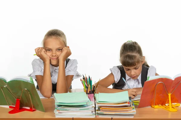 Two schoolgirls at a desk, one leaning on her hands and looking into the frame, the other writes — Stock Photo, Image