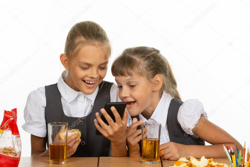 Two schoolgirls at a break watching a video on the phone, and eating liver and orange, drinking juice