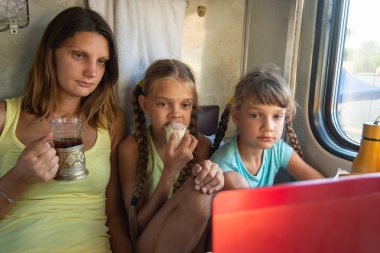 Anne ve iki kız tren vagonundaki dizüstü bilgisayar ekranına bakıyorlar.