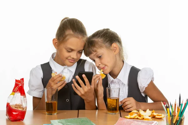 Deux écolières en pause regardent l'écran d'un smartphone, mangent une orange et boivent du jus — Photo