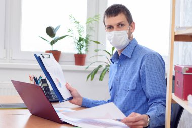 Tıbbi maske takan ofis çalışanı, belgelerdeki verileri şaşkınlıkla inceliyor.
