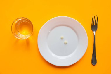 Tabağın üzerinde bir avuç dolusu hap, yanında bir bardak su ve bir çatal var.