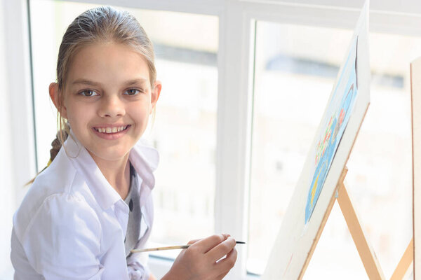 Happy teenager girl draws on an easel by the window and looked into the frame with a smile