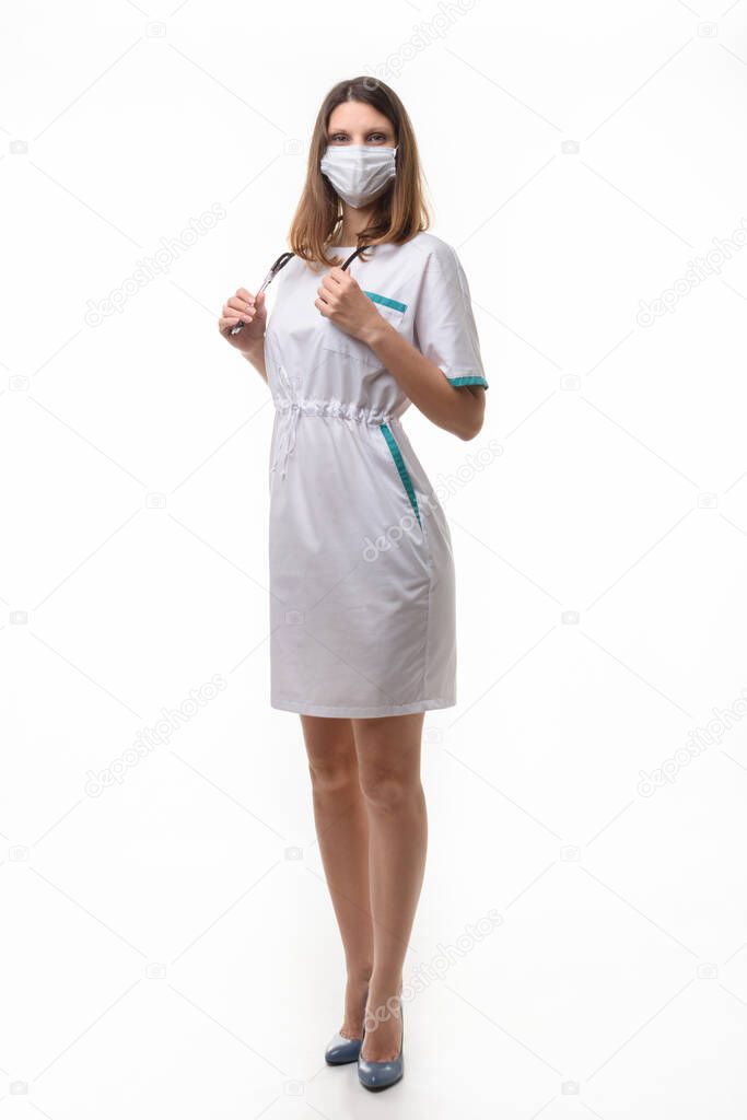 A girl in medical clothes and a mask holds a stethoscope on a white background