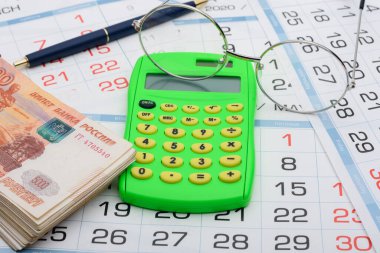 Çerçevenin ortasında hesap makinesi, kenarlarda para, bir kalem ve gözlük.