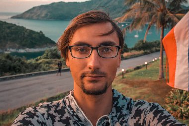 A man with glasses takes a selfie against the backdrop of the sea and mountains Phuket Thailand