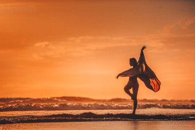 Gün batımında deniz kenarında uçan pelerinle dans eden genç bir kadın.