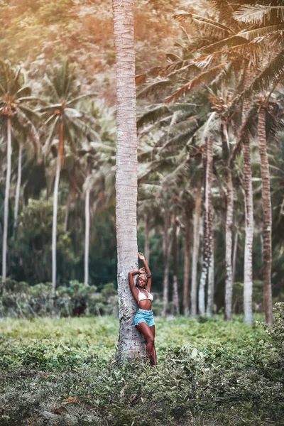 African girl in white bra and blue shorts standing near big palm tree in a sunny palm grove