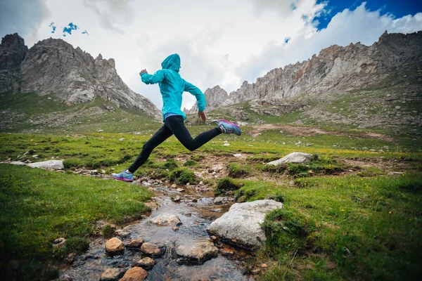 Woman trail runner jumping over small river on beautiful mountains