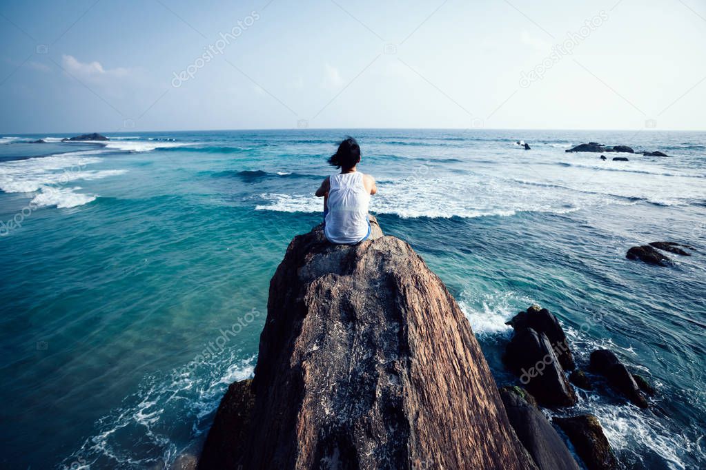Young woman sitting on seaside rock cliff edge and looking at the distance
