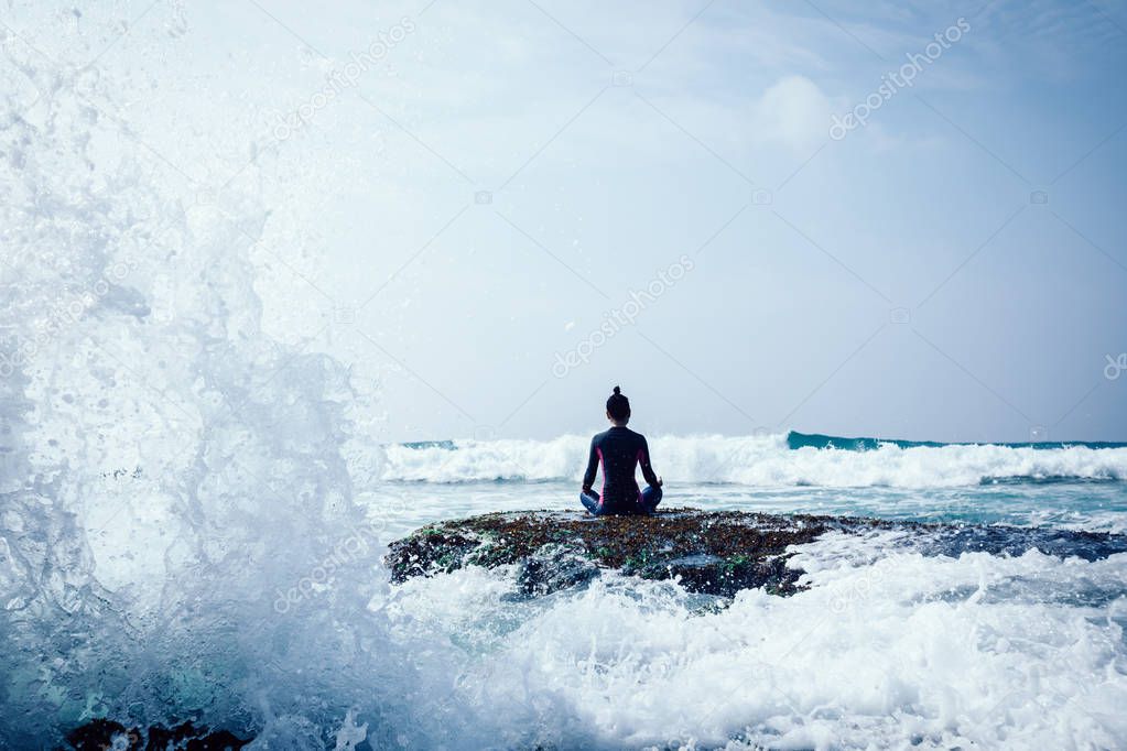Young woman meditating at the seaside cliff edge facing the coming strong sea waves
