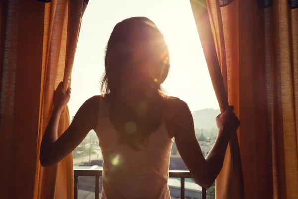 開いているカーテンで部屋に立って 日の出を見て若い女性 — ストック写真