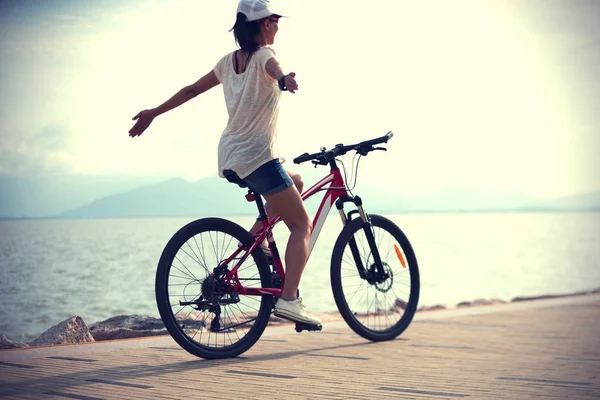 腕を伸ばして日当たりの良い海辺で自転車に乗る女性 — ストック写真