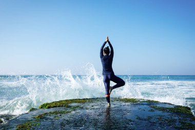 Deniz kenarındaki kayalıklarda yoga yapan kadın güçlü deniz dalgalarıyla karşılaşıyor.
