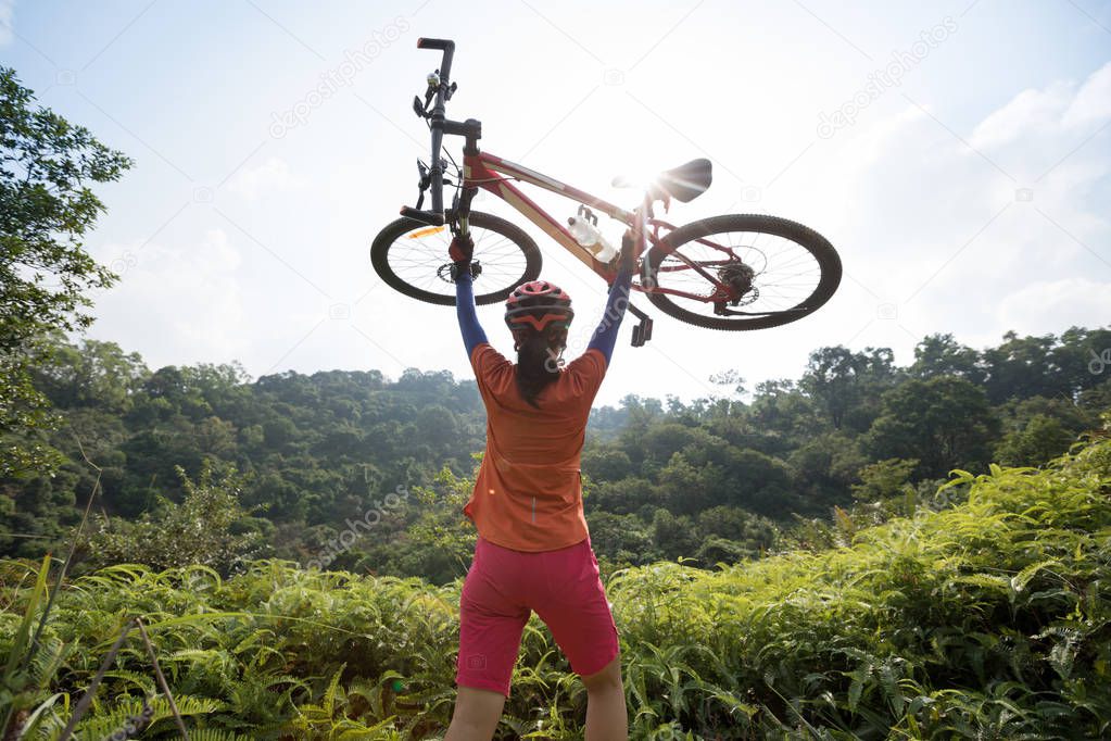 Cross country biking woman cyclist cheering with lifting mountain bike on tropical forest mountain top to sunrise