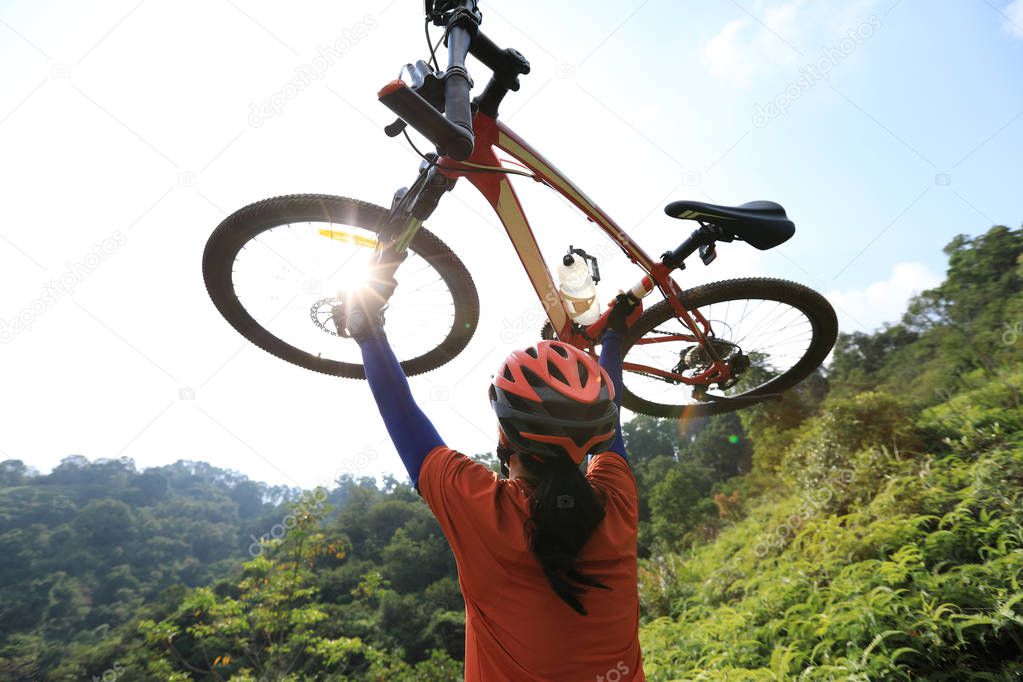 Cross country female cyclist cheering and lifting mountain bike on tropical forest mountain top to sunrise
