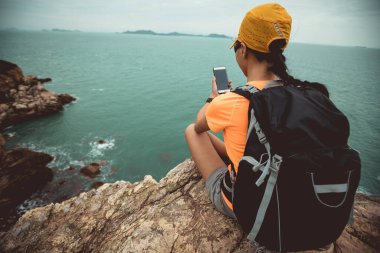 Deniz kenarındaki uçurumun kenarında akıllı telefonla fotoğraf çeken bir kadın yürüyüşçü.