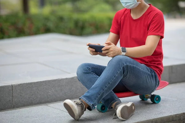 フェイスマスクを身に着けているアジアの女性は 現代都市でスマートフォンを使用してスケートボード上に座る — ストック写真