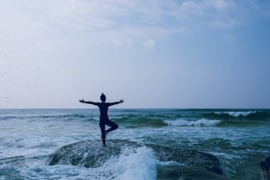 Kadın, yaklaşan güçlü deniz dalgalarıyla yüzleşmek için deniz kenarındaki uçurumun kenarında yoga yapıyor.