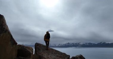 Arka planda karlı dağlar olan, göl kenarındaki kayalıklardaki bir insanın videosu..