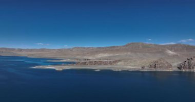 Açık gökyüzünün altındaki kayalık kayalıklarla çevrili derin mavi bir gölün çarpıcı videosu. Tibet, Çin  