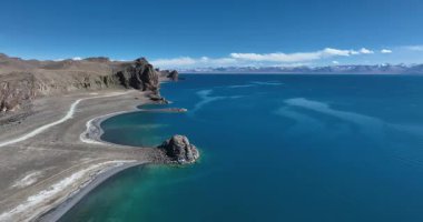 İnsansız hava aracı videosu açık bir gökyüzünün altında derin mavi bir göl, kayalık bir kıyı şeridi ve dağlar gösteriyor..