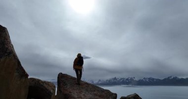 Bir gölün ve karlı dağların kenarındaki büyük kayalıklarda duran birinin arka videosu..