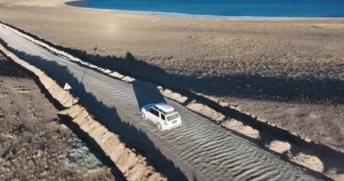 Yüksek rakımlı göl kenarında çalışan beyaz off-road aracının hava görüntüsü.