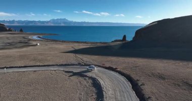 Yüksek rakımlı göl kenarında çalışan beyaz off-road aracının hava görüntüsü.