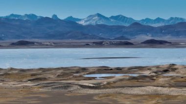 Göl, karlı dağlar ve açık mavi gökyüzü olan geniş, düz bir arazinin havadan çekilmiş videosu..