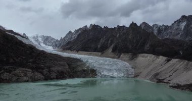 Yüksek açılı bir video, engebeli dağlarla çevrili turkuaz bir göle akan bir buzulu gösteriyor..