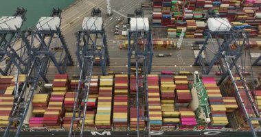 Kalabalık bir ticari limana yanaşan büyük bir konteynır gemisinin arkasında yığınla kargo konteynırı, dağ ve şehir vardı..