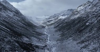 Bulutlu, bulutlu bir gökyüzünün altında kayalık tepelerle çevrili bir dağ vadisinde akan büyük bir buzulun dramatik bir görüntüsü..