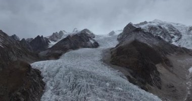 Bulutlu, bulutlu bir gökyüzünün altında kayalık tepelerle çevrili bir dağ vadisinde akan büyük bir buzulun dramatik bir görüntüsü..