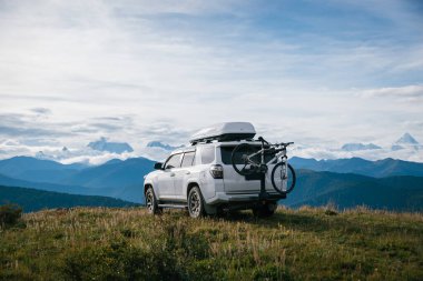 Dağların arka planında siyah sırt çantalı beyaz bir SUV..