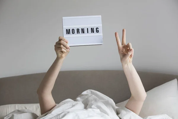 Female in bed under the sheets holding up a morning sign — Stock Photo, Image