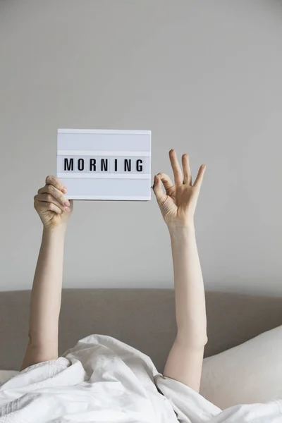 Γυναίκα στο κρεβάτι κάτω από τα σεντόνια κρατώντας ένα πρωινό σημάδι — Φωτογραφία Αρχείου