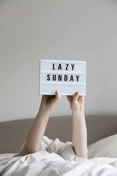 Female in bed under the sheets holding up a lazy sunday sign — Stock Photo, Image
