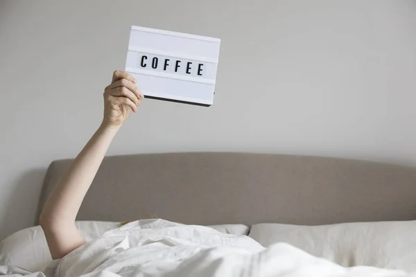 Mujer en la cama debajo de las sábanas sosteniendo un cartel de café — Foto de Stock