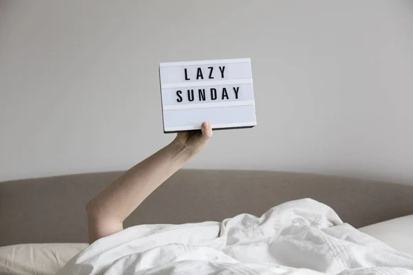 Female in bed under the sheets holding up a lazy sunday sign — Stock Photo, Image