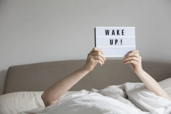 Female in bed under the sheets holding up a wake up sign — Stock Photo, Image