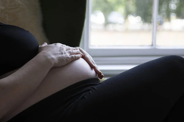 Close up of a prgnant woman holding her baby bump while relaxing on a chair — стоковое фото