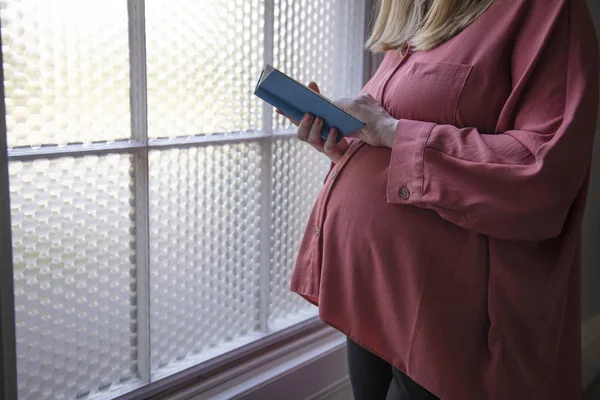 Una donna prgnant che legge una bibbia blu vicino ad una finestra luminosa — Foto Stock